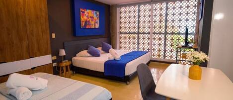 Habitación de hotel con dos camas, una mesa de comedor, una mesita con un florero y un cuadro de colores en la pared.
