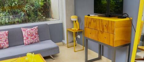 Una sala moderna con un sofá, una mesa de centro amarilla, un televisor sobre un mueble de madera y una ventana con vista a follaje.