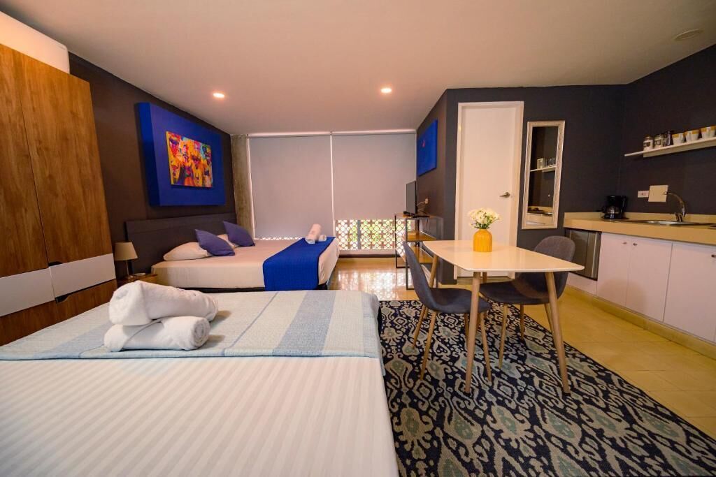 A hotel room with a bed, a desk, a chair, a small table with a vase, a kitchenette, and a large window.