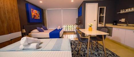 A hotel room with a bed, a desk, a chair, a small table with a vase, a kitchenette, and a large window.