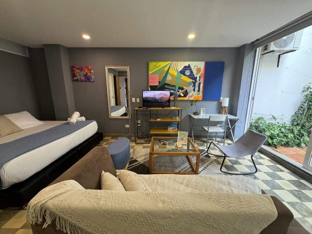 Habitación de hotel moderna con cama, sofá, escritorio y silla. Hay un cuadro en la pared y una ventana con vistas a zonas verdes.