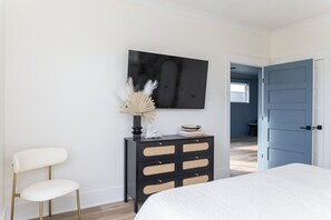 Ein Schlafzimmer mit einem Bett, einer schwarzen Kommode mit Schubladen, einem weißen Stuhl und einem an der Wand befestigten Fernseher.