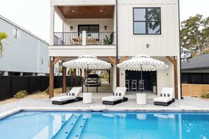 Une maison à deux étages avec un espace piscine comprenant des chaises longues et des parasols.