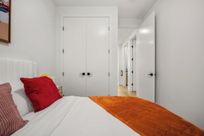A bedroom with a bed, white walls, a white door, and a hallway leading to another room. - BEST Stylish 3BR | WFH Space | 7 mins to DT (Calgary)