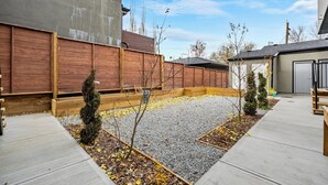 Un jardin à l’arrière d’une maison, avec une terrasse en béton, une clôture en bois et quelques petits arbres.