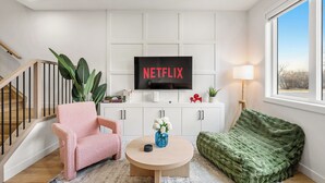 Un salon avec un téléviseur affichant le logo « NETFLIX », un fauteuil rose, un canapé vert et une table basse en bois.