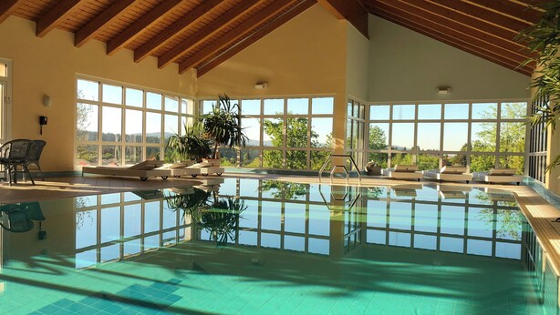 Ein Innenpool mit klarem, blauem Wasser, Holzbalken an der Decke und großen Fenstern mit Blick auf die grüne Landschaft.