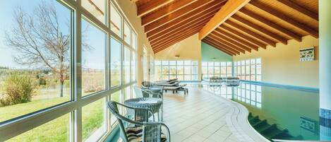 An indoor swimming pool with clear views of the outdoors, surrounded by large windows and furnished with lounge chairs.