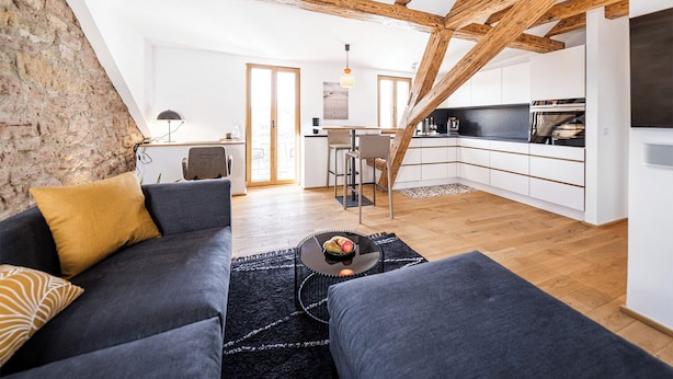 A modern loft apartment with exposed wooden beams, a cozy seating area with a sofa and a coffee table, a kitchen with white cabinetry, and a large window providing natural light. - Stylish apartment in Tiefenbach (Östringen)