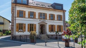 A two-story building with a sign reading "ALTE SCHULE." It features a traditional facade with wooden shutters, a central entrance, and a small garden area with potted plants. - Stylish apartment in Tiefenbach (Östringen)