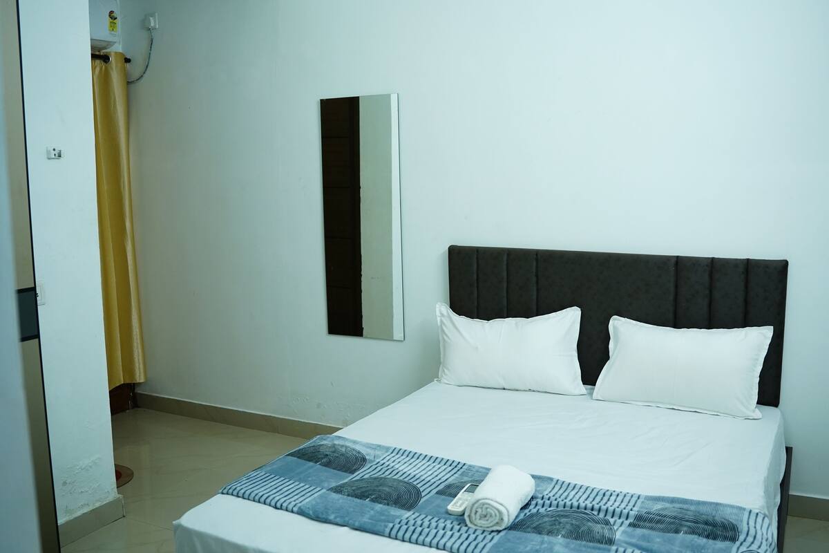 A neatly made bed with a patterned bedspread and two pillows, a bedside mirror, and a door with a curtain partially visible.