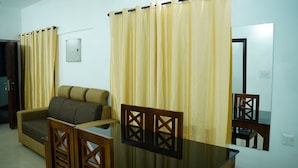 A room with a sofa, a glass-top table, wooden chairs, and curtains. - Stay Ease Rental Rooms (Palakkad)