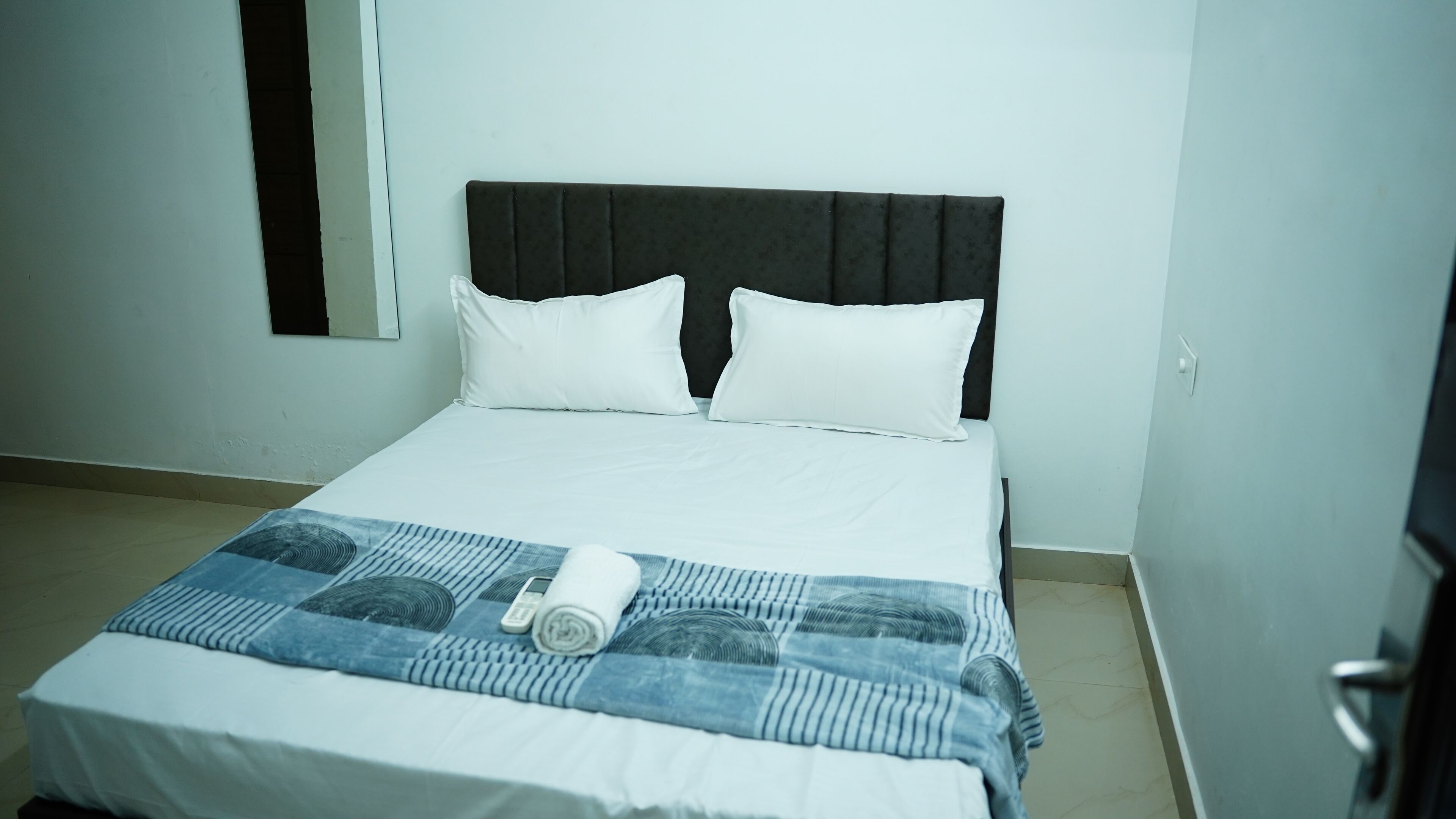 A neatly made bed with white bedding and a blue patterned blanket, a dark headboard, and a small window with a black frame.