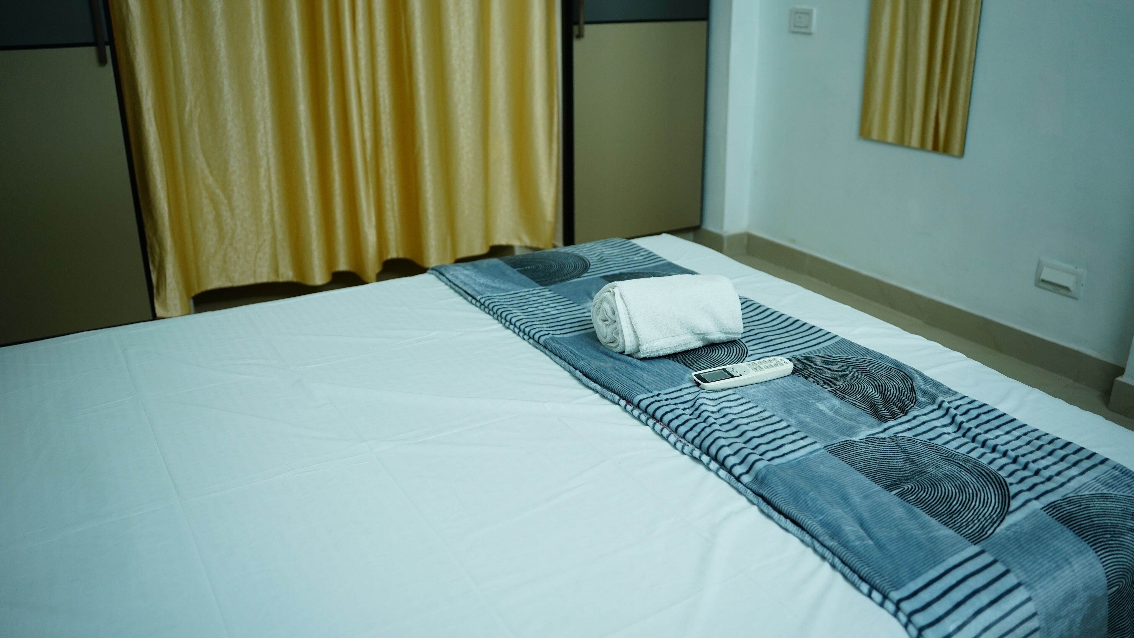 A neatly made bed with a white sheet, a rolled white towel, and a remote control on a blue and white patterned blanket.