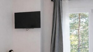 Ein Zimmer mit Schreibtisch aus Holz, einem Sessel, einem flachbildfernseher an der Wand und einem Fenster mit Blick auf Bäume.