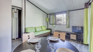 Huoneessa on ruokapöytä, jonka ääreen on katettu neljälle, vihreä sohva, televisio ja ikkuna, jossa on verhot.