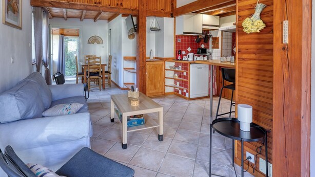 Ein gemütliches Wohnzimmer mit einer Couch, einem Couchtisch und einem Essbereich mit Stühlen. Im Hintergrund ist eine Küche mit Holzschränken und einer Spüle erkennbar.