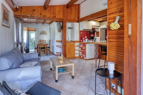 A cozy living room with a sofa, a coffee table, and a dining area with chairs. A kitchen with wooden cabinets and a sink is visible in the background.