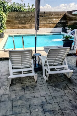 Ett poolområde med två vita solstolar och ett litet bord, omgivet av ett trästaket och krukväxter.