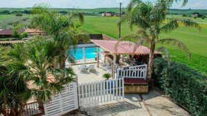 A pool area with a covered seating area, surrounded by palm trees and a white fence. - Villa Lucia (Lucera)