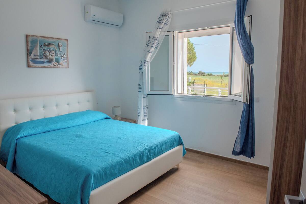 A bedroom with a bed, a window with curtains, a painting on the wall, and a view of the outdoors.