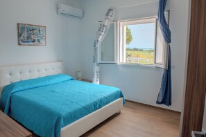 Un dormitori amb llit, finestra amb cortines, un quadre a la paret i vista a l'exterior.