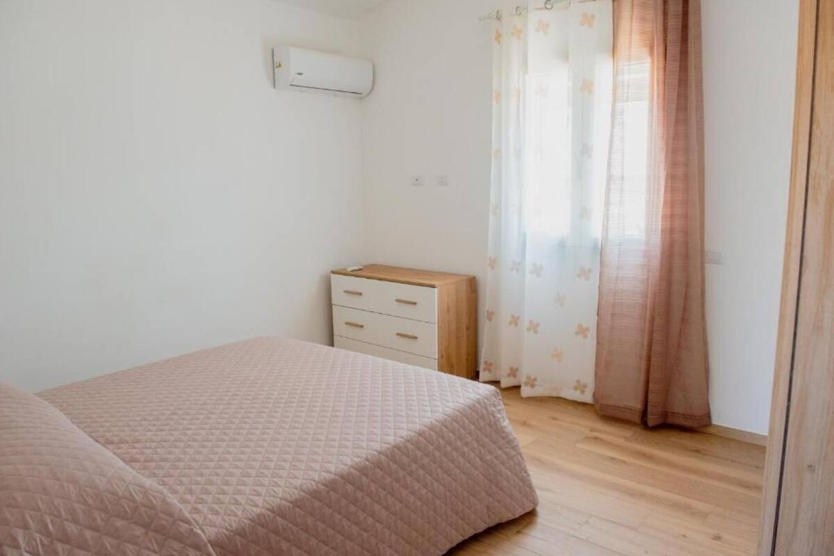 A bedroom with a bed, a small bedside table, a drawer unit, and a window with curtains.