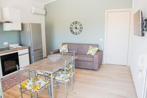 A compact living space with a kitchenette, a dining table with floral chairs, a sofa with matching cushions, and a wall clock. - Villa Rosy (Butera)