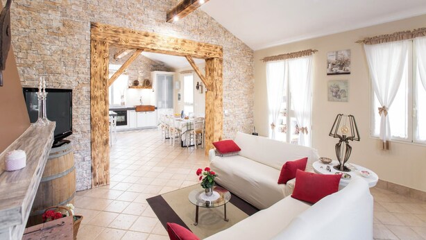 A spacious living room with a stone wall, wooden beams, a white sofa with red cushions, a TV, and a dining area in the background. - Villetta Tra Mari E Monti (San Giovanni Rotondo)