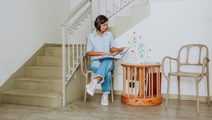 Une personne est assise sur une chaise près d’un escalier, tenant un livre. Une petite table ronde et une autre chaise se trouvent à proximité.