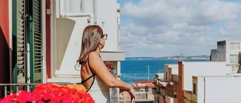 A balcony with a view of the sea, a building with green shutters, and a person leaning on the railing.