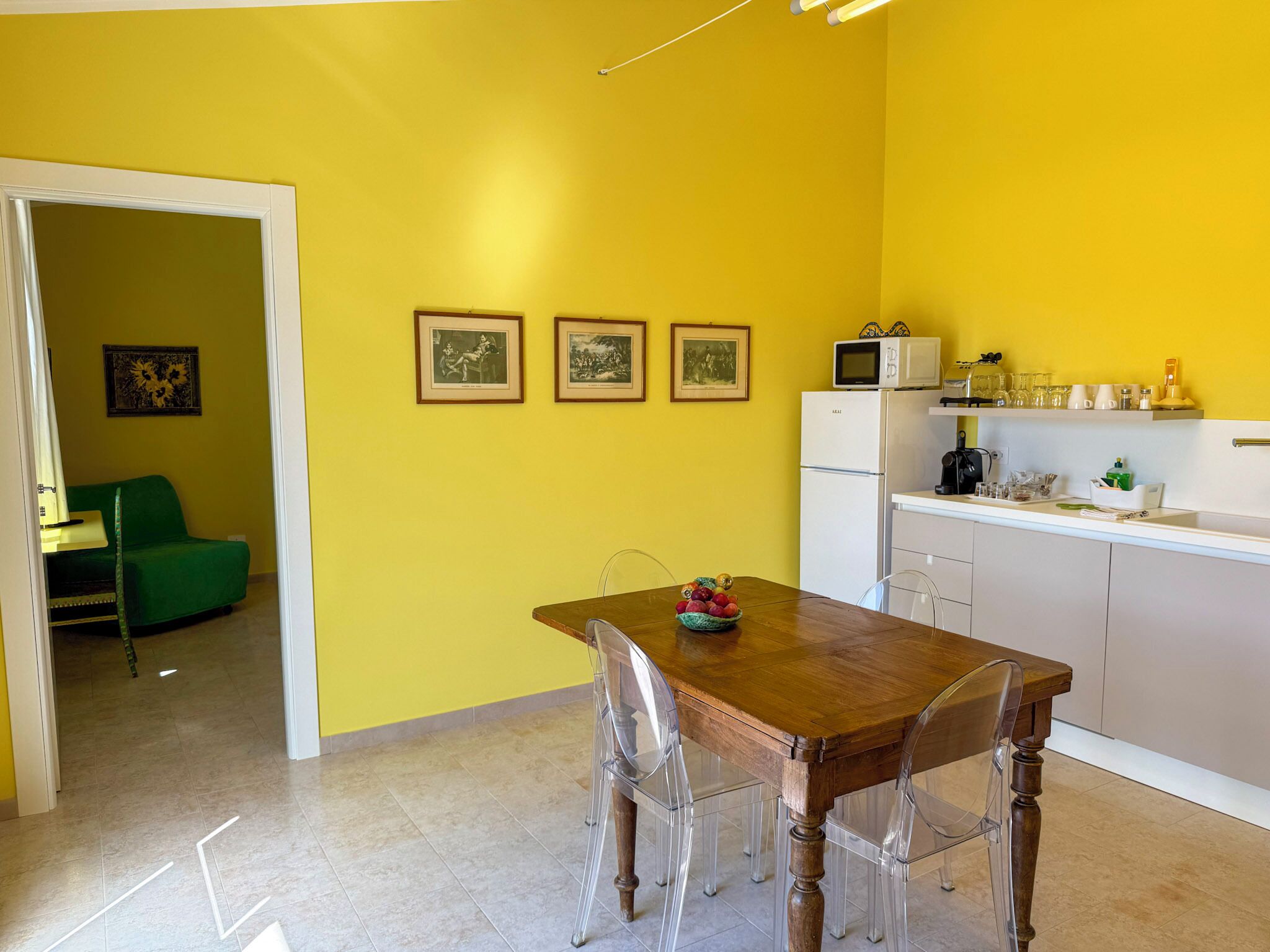 A compact kitchen with a wooden table and chairs, a white refrigerator, and a microwave. There are three framed pictures on the wall.
