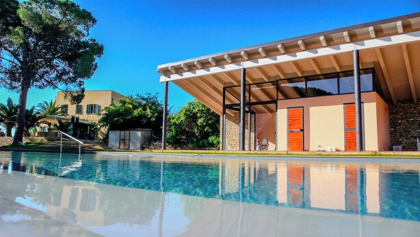 A modern building with a pool in front, featuring a clear blue sky and surrounding greenery.