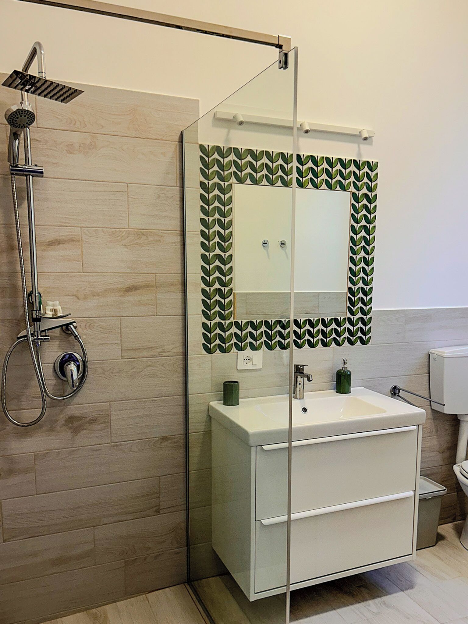 A modern bathroom with a glass shower enclosure, a white sink with a green soap dispenser, and a toilet.