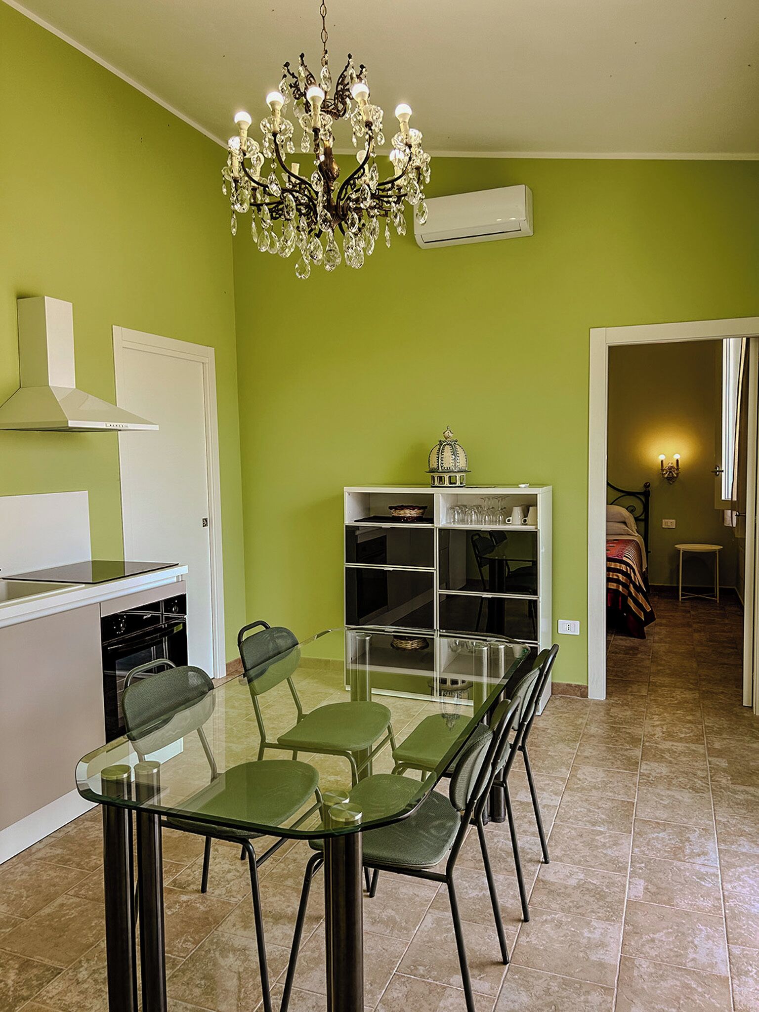 A dining area with a glass table and chairs, a chandelier, a kitchen with a stove and oven, and a bookshelf.