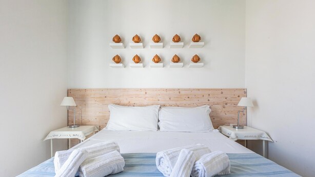 A neatly made bed with white linens and towels, a wooden headboard, and a wall-mounted shelf with decorative items. - L`Uomo e il Mare - Camera Vista Mare (Gallipoli)