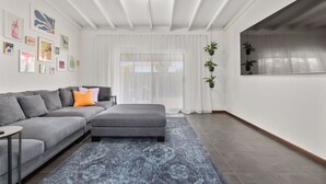 Ein modernes Wohnzimmer mit einer grauen Eckcouch, einem kleinen Beistelltisch, einem gemusterten Teppich und einem großen Fenster mit transparenten Vorhängen.