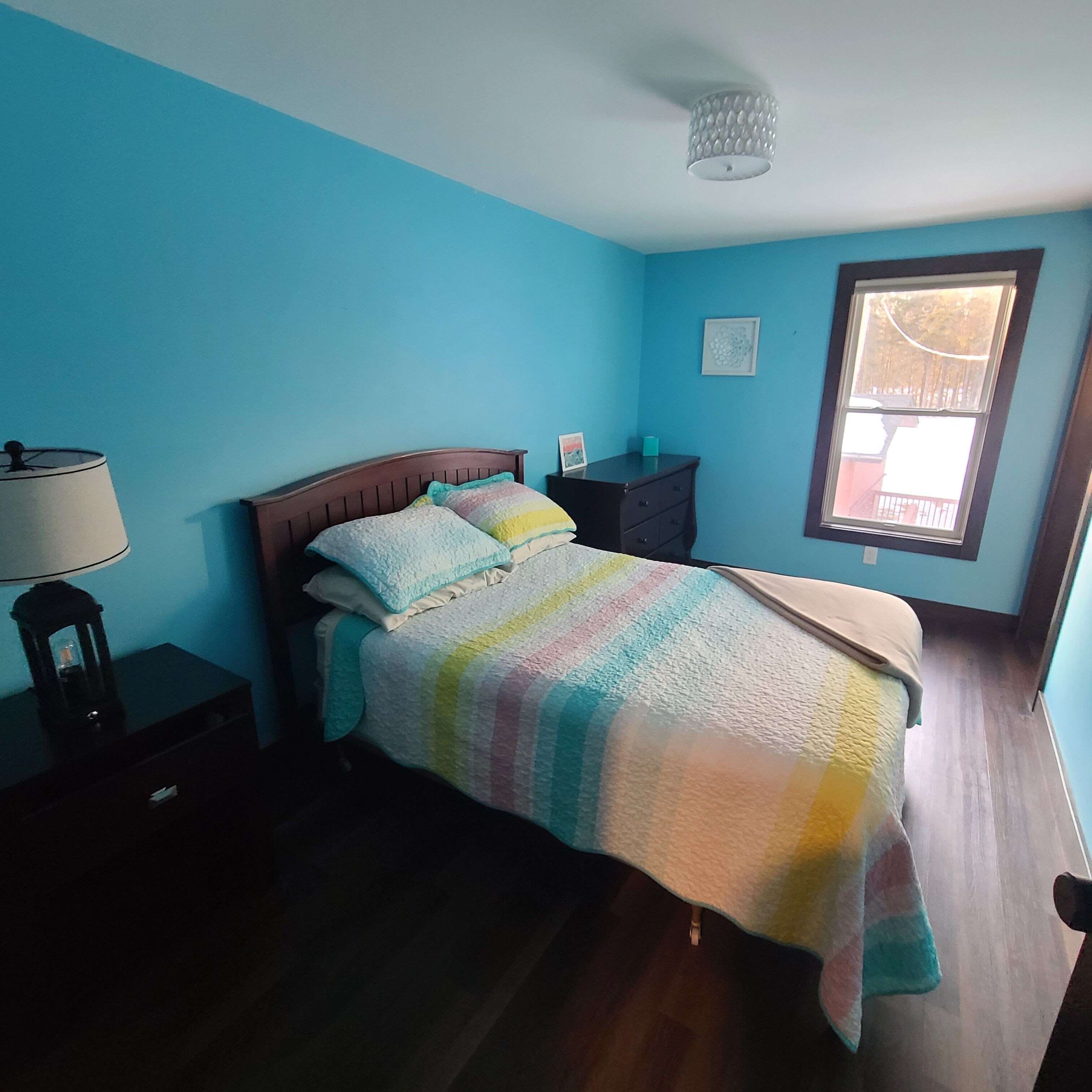 A bedroom with a bed, a nightstand, a lamp, a dresser, and a window with a view.