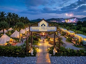 En resortindgang med et skilt for Wisdom Village Pattaya, der viser en sti til hovedområdet, omgivet af telte og palmer.
