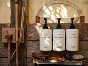 Un estante de madera con tres botellas blancas etiquetadas 'BYREDO BAL D'AFRIQUE' para champú, acondicionador y jabón de cuerpo. También hay un peine marrón y una toalla enrollada sobre el estante.