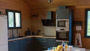 Interior de una cabaña de madera con una mesa de comedor, sillas, una zona de cocina con electrodomésticos y una lámpara colgante.