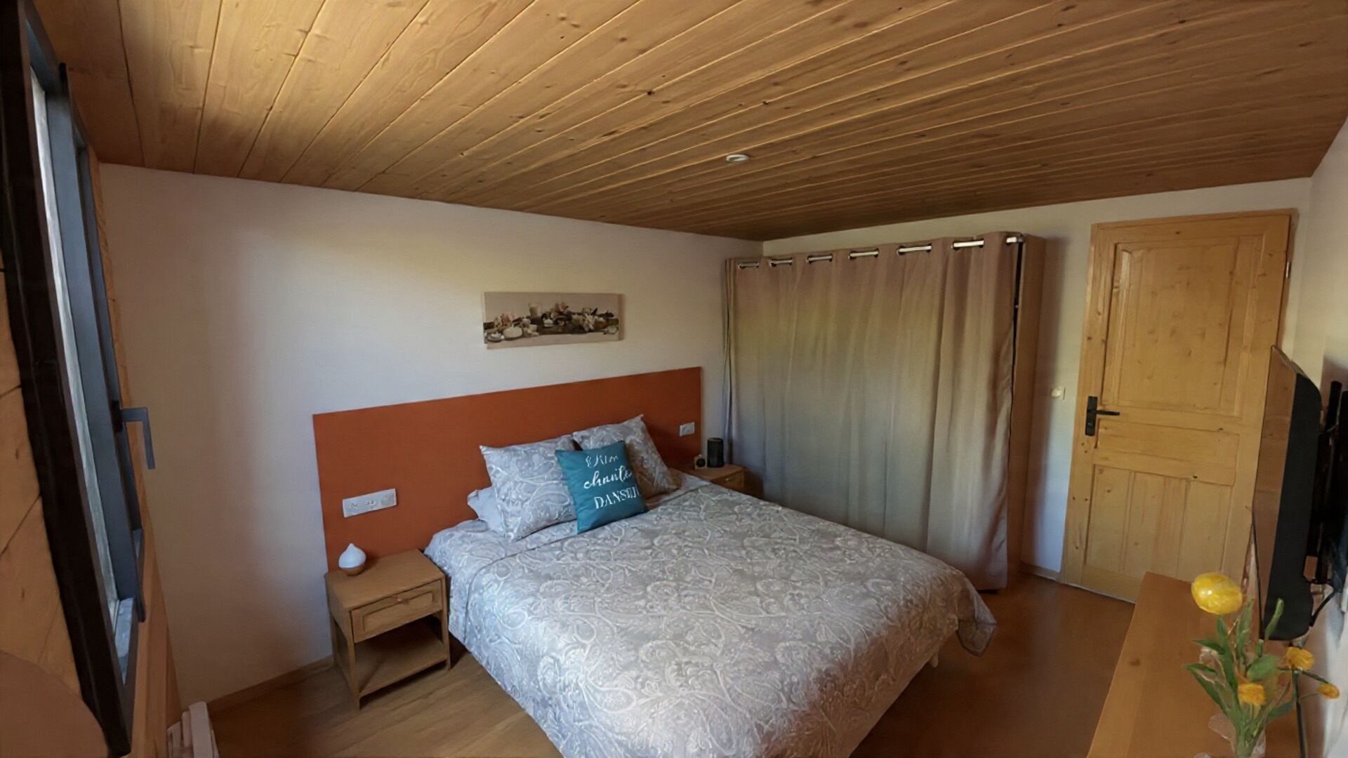 Une chambre avec un plafond en bois, un lit avec une literie à motifs, une table de chevet, une lampe fixée au mur et une porte.