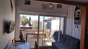 A cozy living room with a sofa, a table with chairs, a TV, and a sliding glass door leading to a patio with a bicycle and a wooden fence. - Duplex Apartment for a Beach Vacation (LE GRAU D'AGDE)