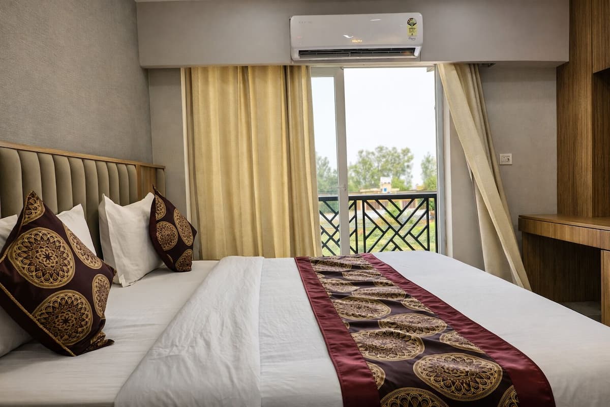 Habitación de hotel con una cama grande, cabecero, ventana con cortinas, unidad de aire acondicionado y balcón con barandilla.