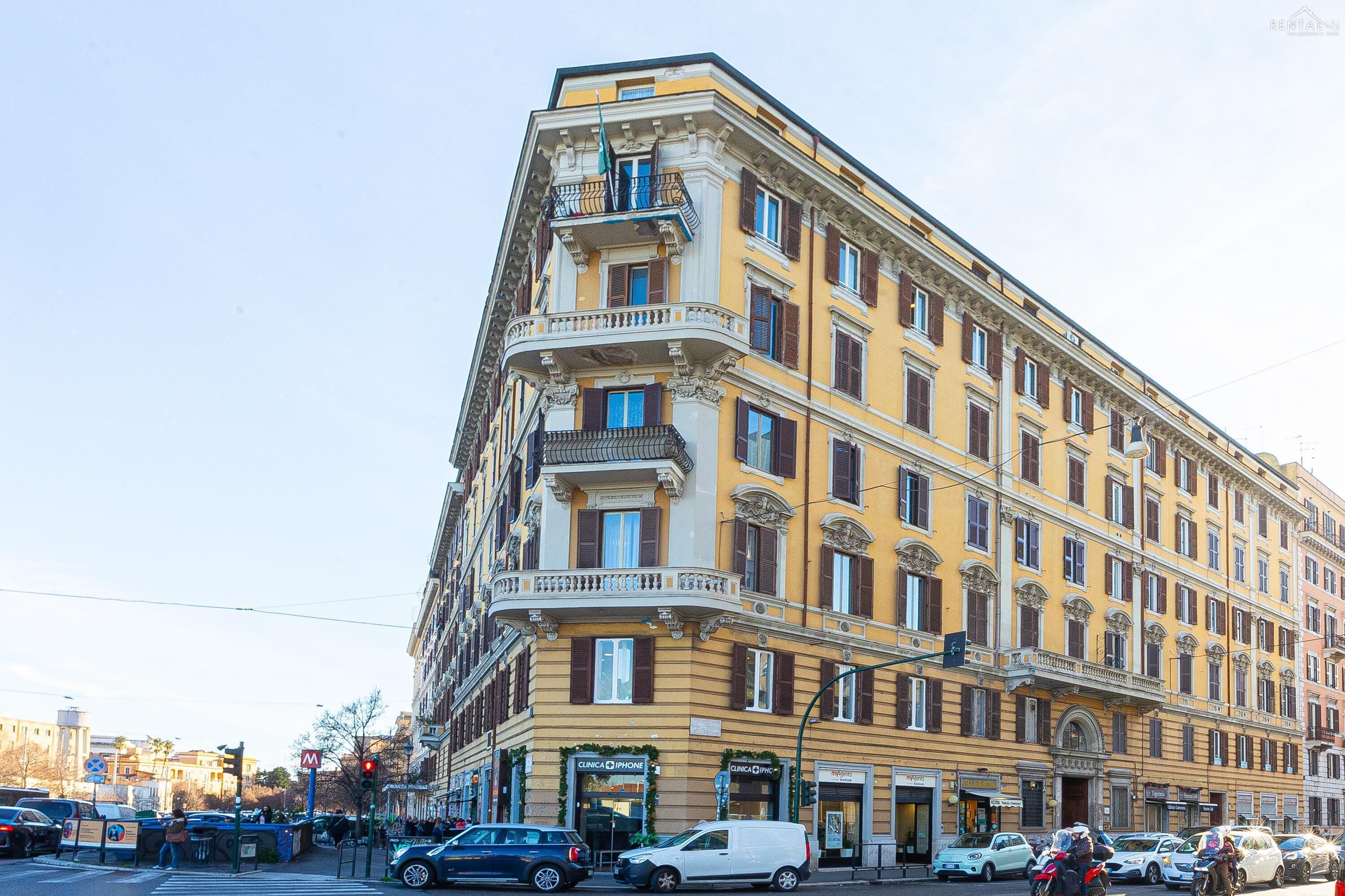 A multi-story building with balconies, a clinic on the ground floor, and parked cars along the street.