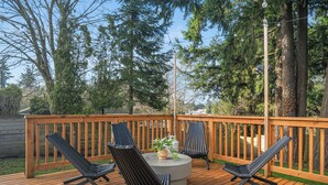 Eine Holzterrasse mit zwei Liegestühlen und einem runden Tisch, umgeben von einem Holzgeländer und Bäumen.