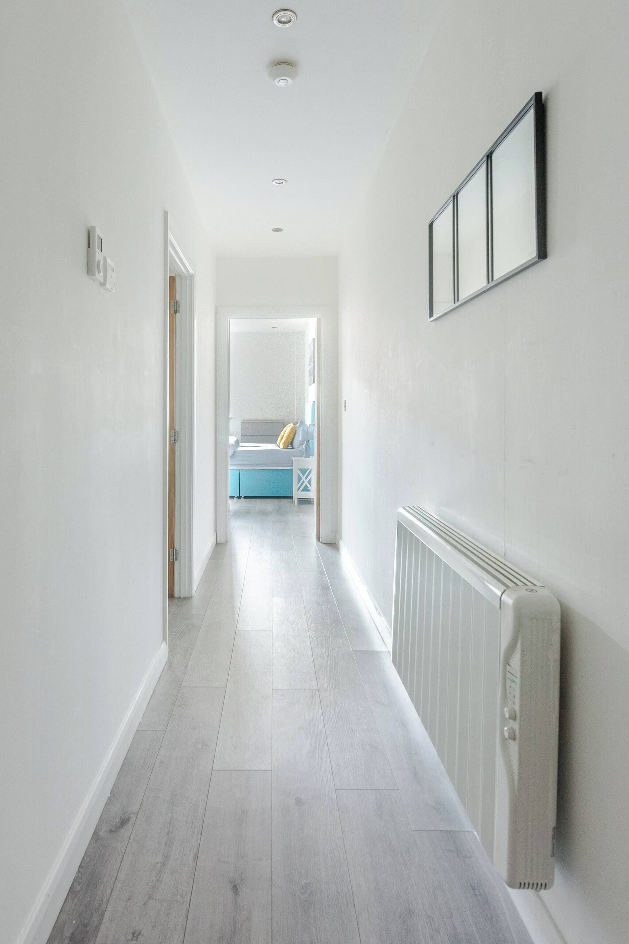 A hallway with a radiator, a framed picture on the wall, and a bed with a blue headboard in the background.