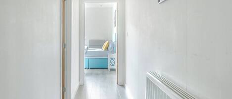 A hallway with a radiator, a framed picture on the wall, and a bed with a blue headboard in the background.