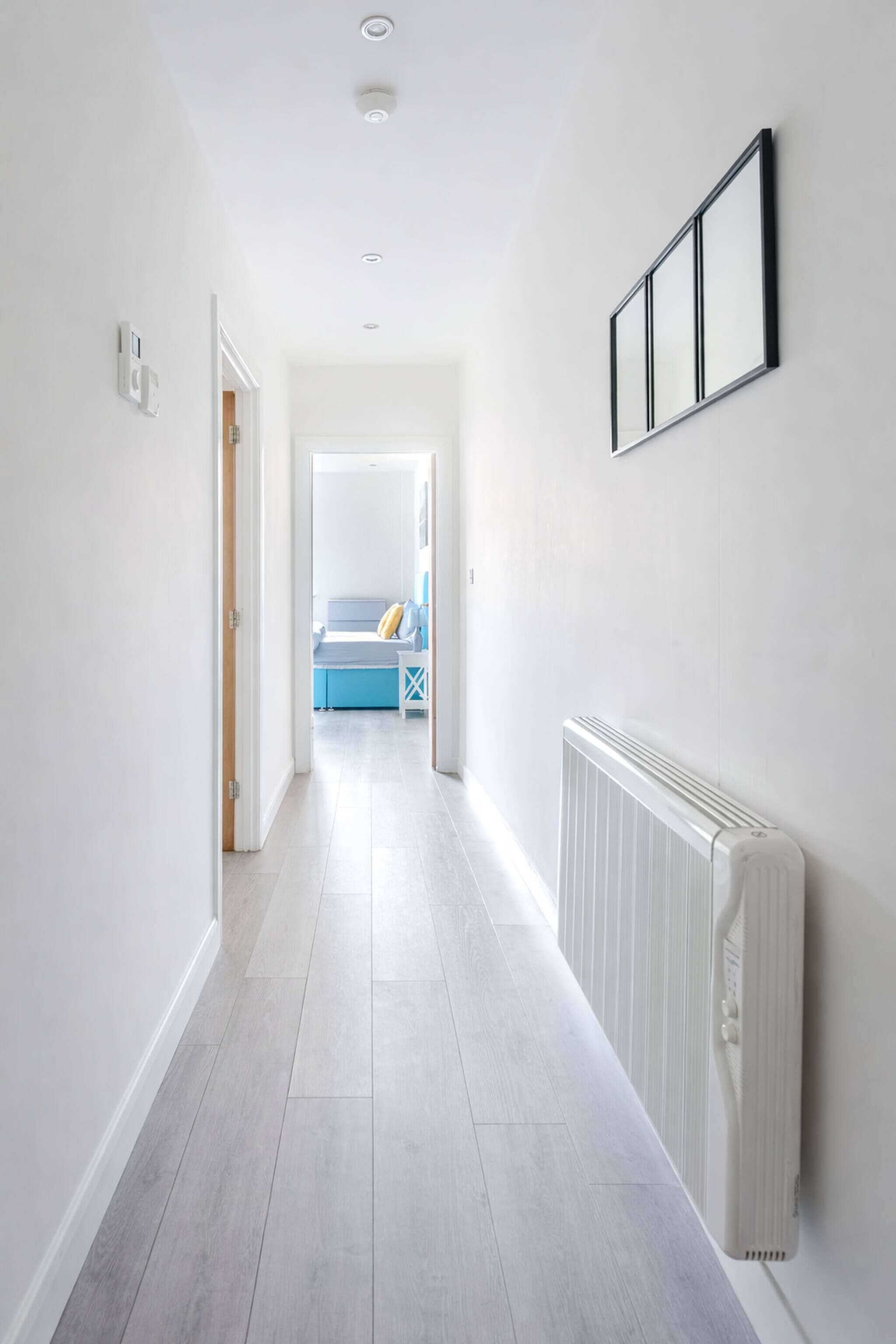 Un couloir avec un radiateur, un tableau encadré et une porte qui donne sur une pièce où se trouvent un lit et un canapé bleu.