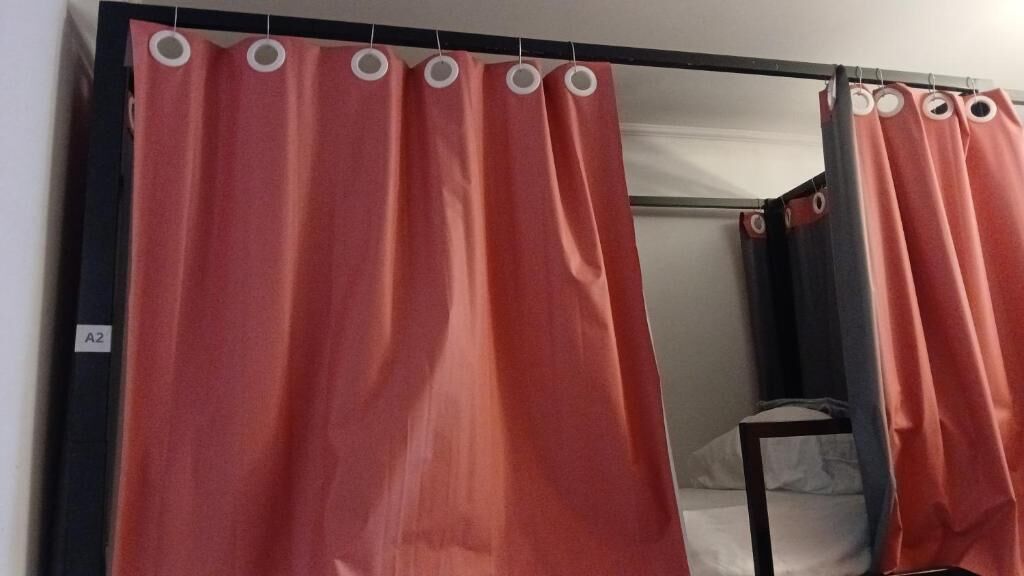 A room with two beds, each with a white mattress and a black frame. There are red curtains with white rings on the windows.